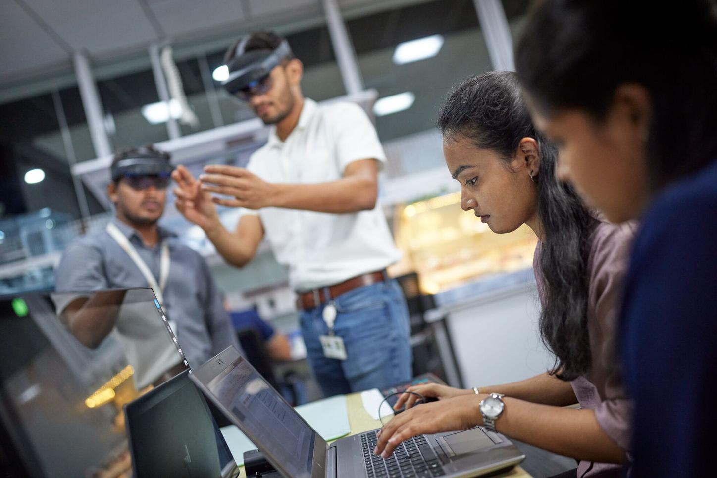 Siemens Healthineers employees from India are working. Two female employees are sitting in front of a laptop, one is typing. Behind them are two male employees with VR glasses on their heads.