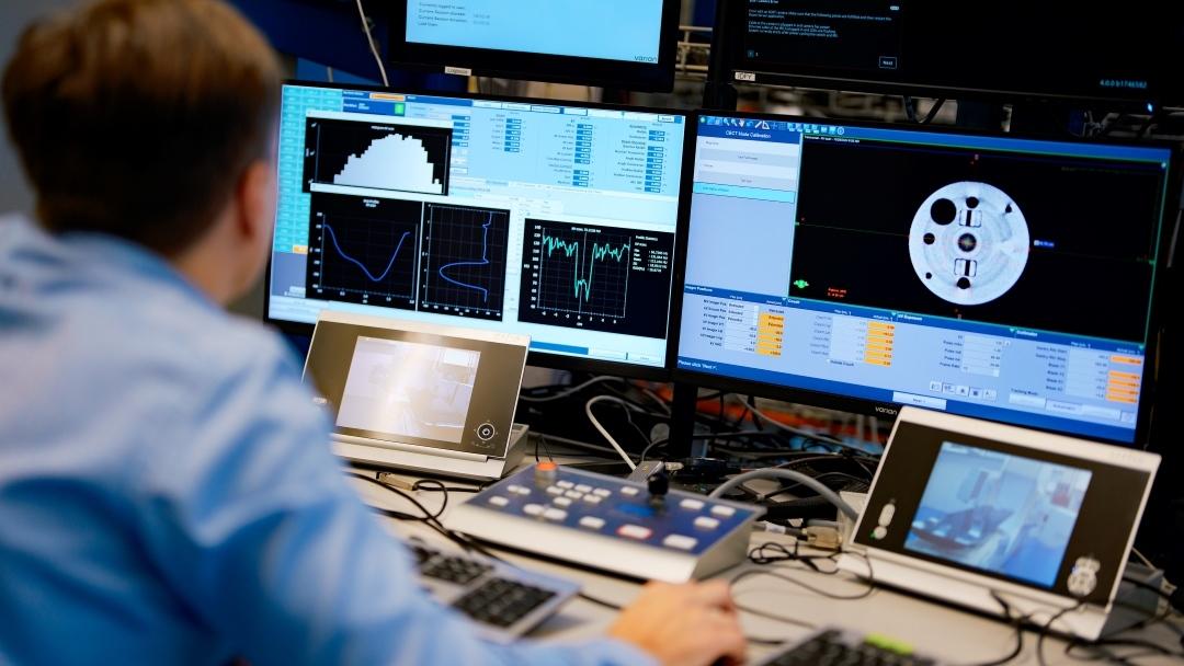 Over-shoulder view of Adam Strzelecki looking at two screens that show scaned images and graphs. There are also two smaller screens at the desk that show the lab room with the radiotherapy device and a control system.