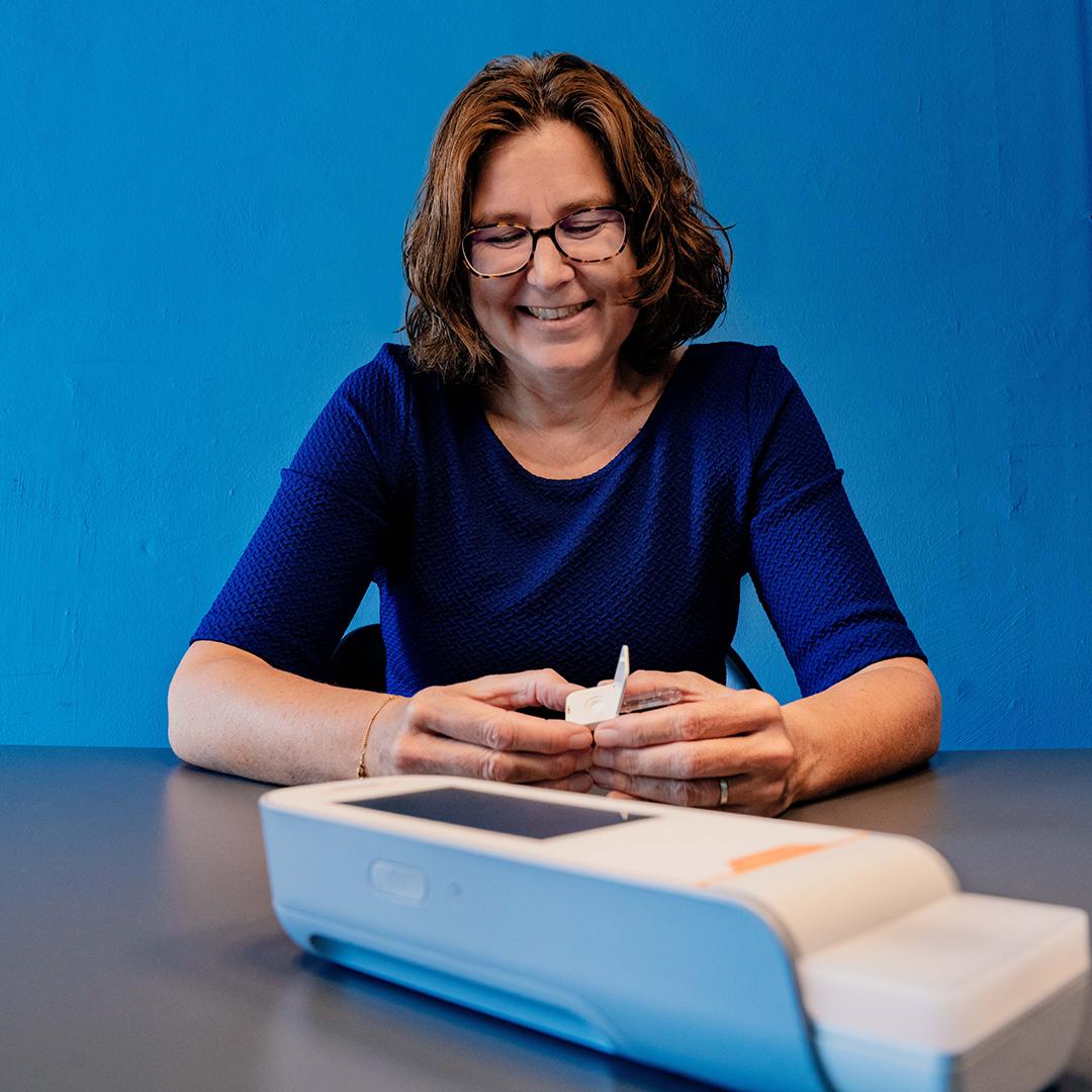 Femke de Theije holding sitting at a desk, holding an open cartridge in her hands, smiling. In the foreground you see the handheld device, Atellica VTLi. In the back is a blue wall.