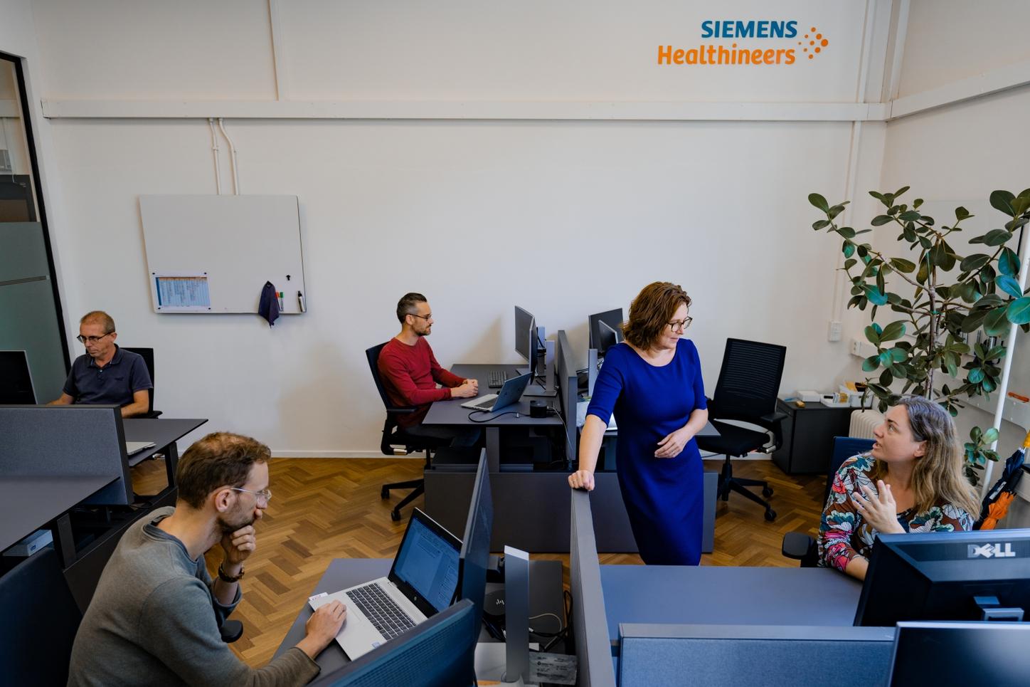 Femke standing in the office, talking to her colleague who's sitting at her desk. Other colleagues are working on their laptops in the back.