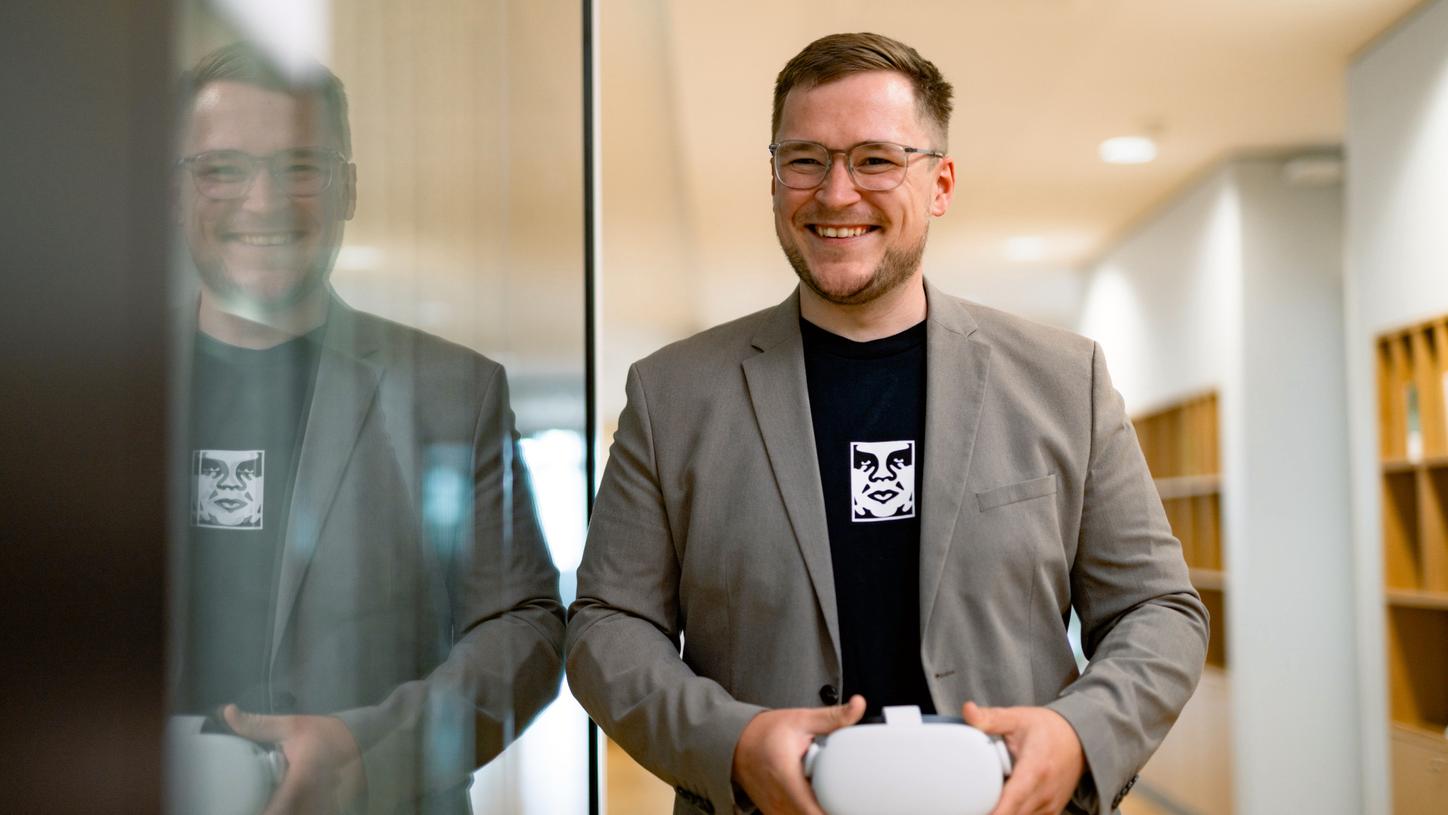 Portrait of Anton. He is smiling, leaning against a glass door, and holding a pair of virtual reality glasses in his hands. You can see his reflection in the glass door.