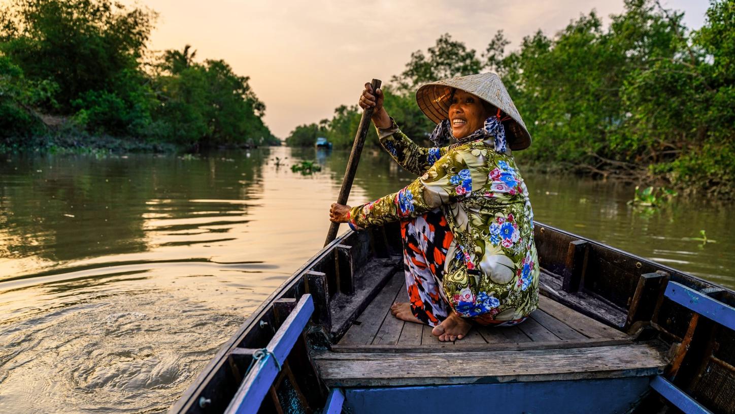 Smiling woman paddling a wooden boat through a river at sunset.