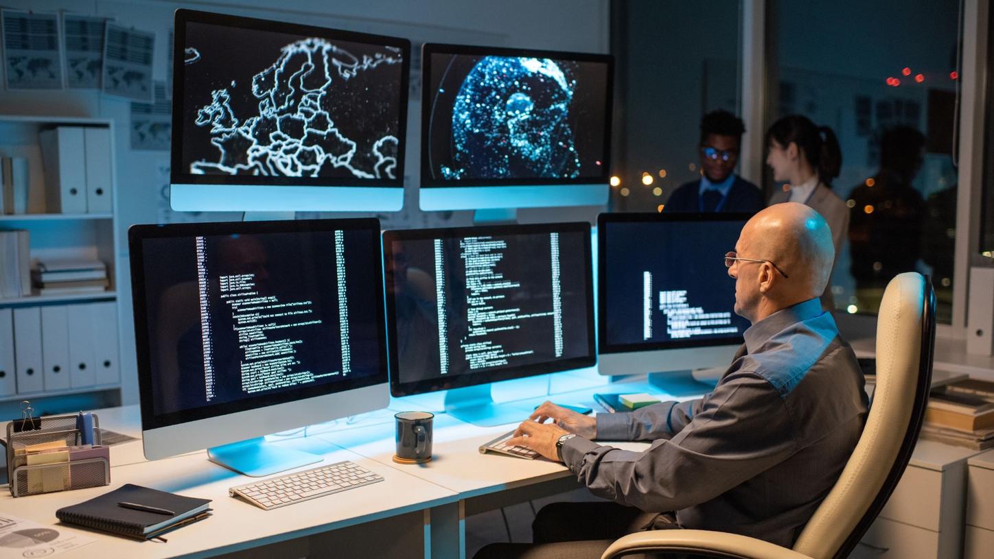 A cybersecurity professional monitors multiple screens displaying code and digital maps.