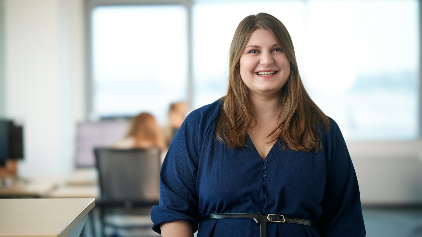 A smiling woman in a navy blue dress stands confidently in a modern office with colleagues working in the background.