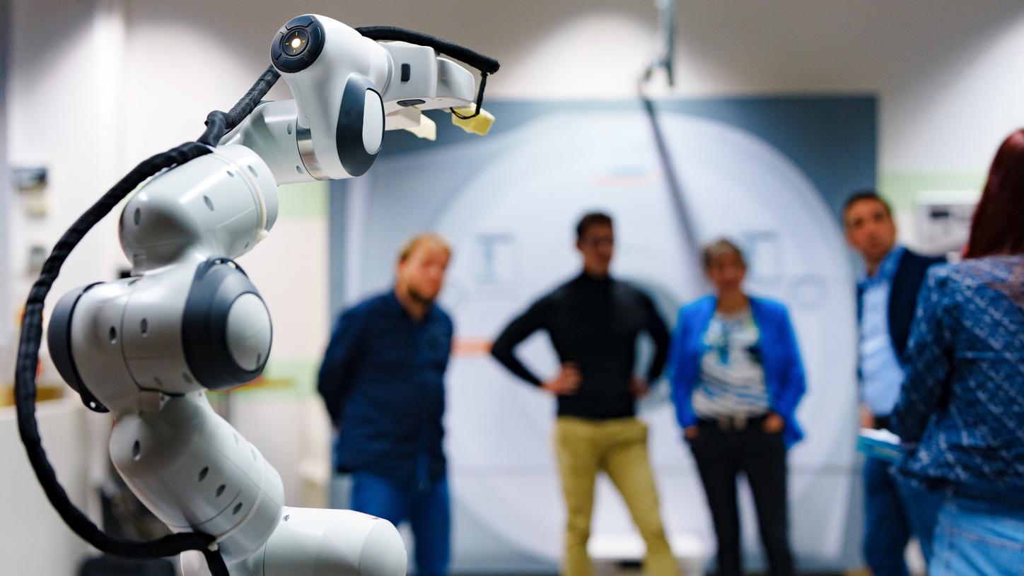 Foto aus dem Innovationslabor. Im Vordergrund sieht man scharf den Greifarm des Prototyps des AURORA-Assistenzroboters. Im Hintergrund stehen Chiheb Dahmani und drei Teammitglieder. Sie sind nur unscharf zu erkennen. 