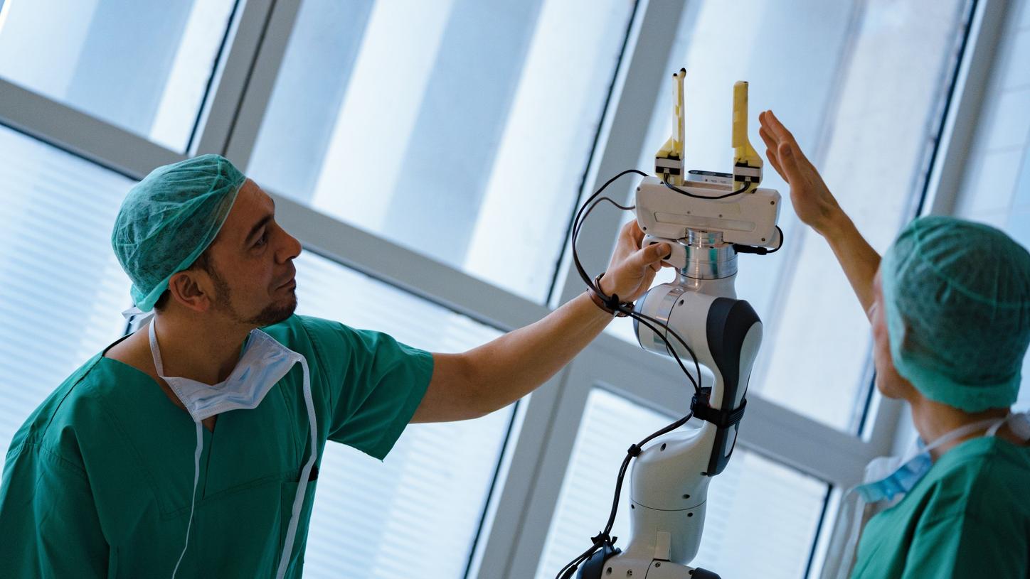 Photo of Chiheb Dahmani and Lukas Bernhard, scientific director of the research group for minimally invasive interdisciplinary therapeutic intervention at the Technical University of Munich hospital. Both men are wearing surgical attire and touching the gripper of the AURORA assistant robot prototype with their hands.