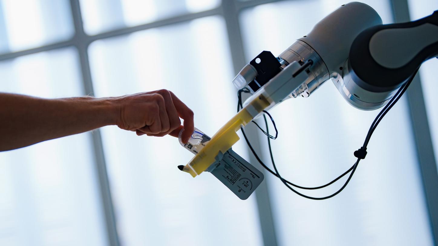 Nahaufnahme. Der Assistenzroboter-Prototyp AURORA übergibt einer menschlichen Hand ein chirurgisches Nahtset. Im Hintergrund ist eine unscharf eine Fensterfront zu erahnen.  