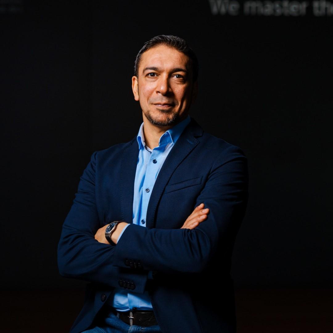 Porträtfoto von Chiheb Dahmani. Er steht vor einem dunklen Hintergrund und lächelt mit verschränkten Armen leicht in Richtung Kamera.