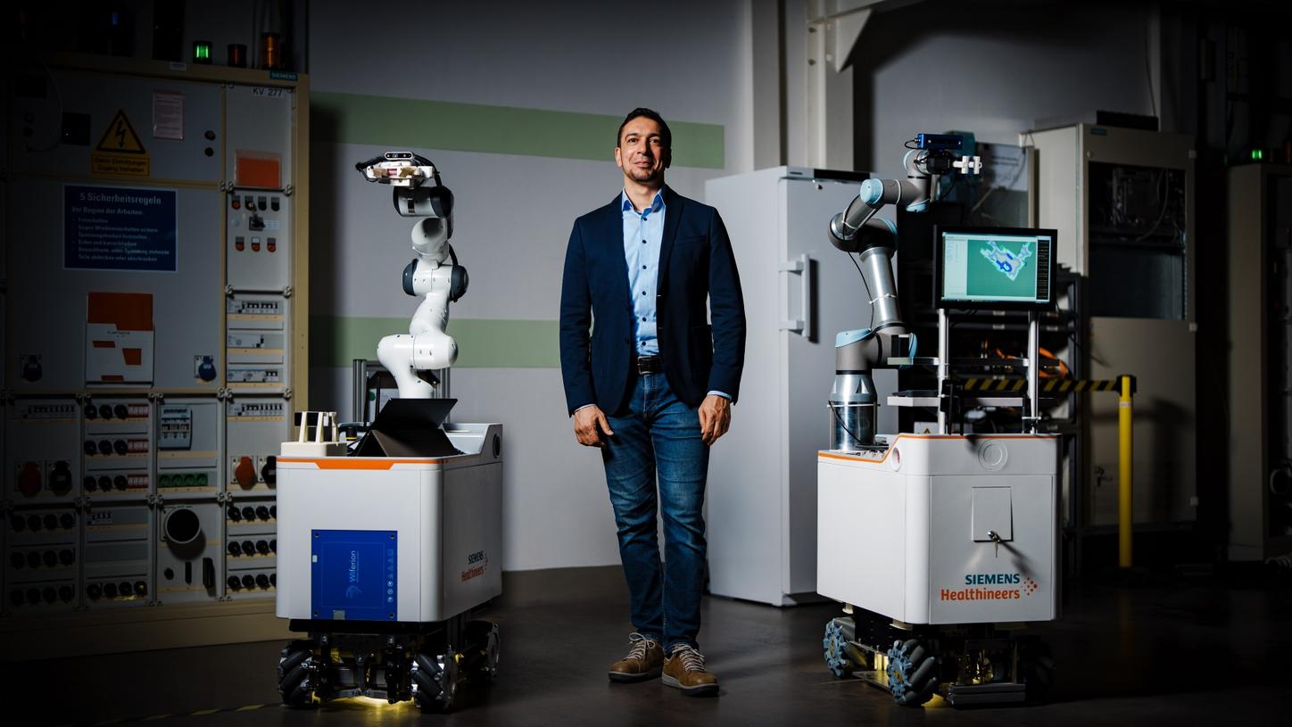 Chiheb Dahmani stands in the innovation lab between the two robot prototypes Vanessa and AURORA and looks at the camera with a slight smile.