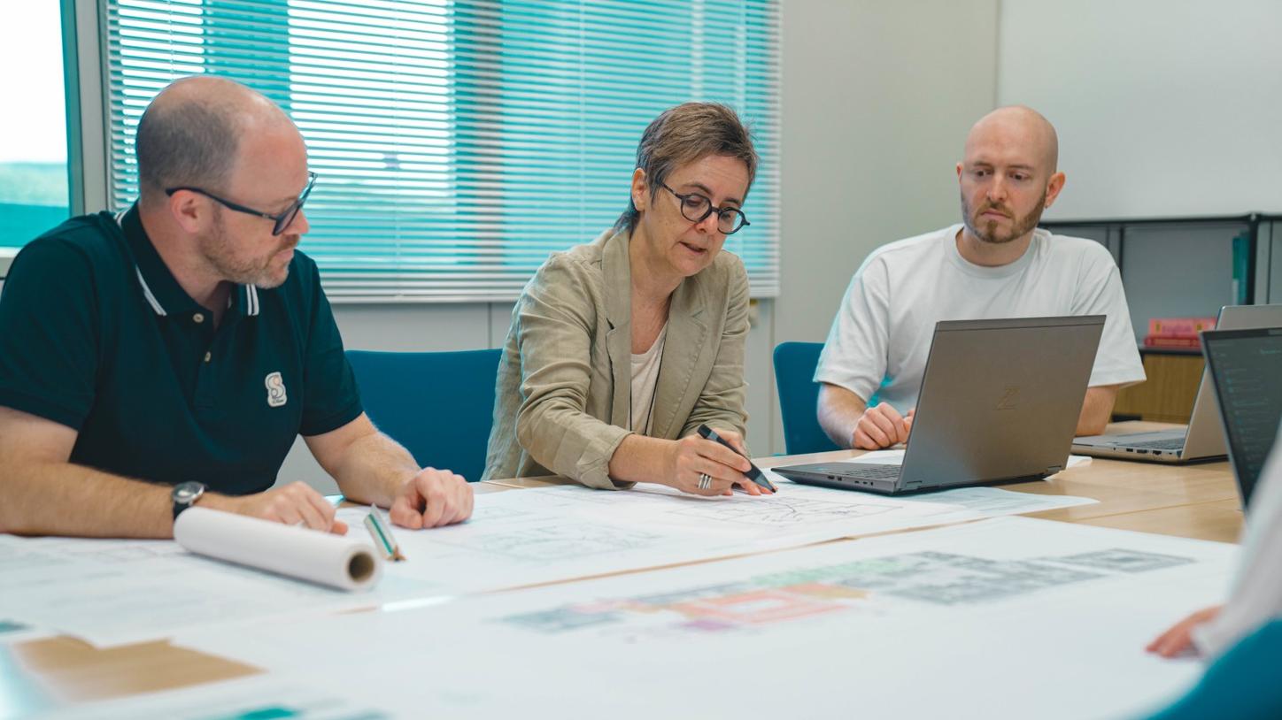 Stefanie Vonhoff is sitting in a conference room in the middle between her colleagues, IT and 3D expert Matthias Brenner and architect Daniel Brehm. On the table is a roll of paper with an architectural plan on which Stefanie is drawing something. Her colleagues are listening attentively. In the foreground you can see more large-format plans.