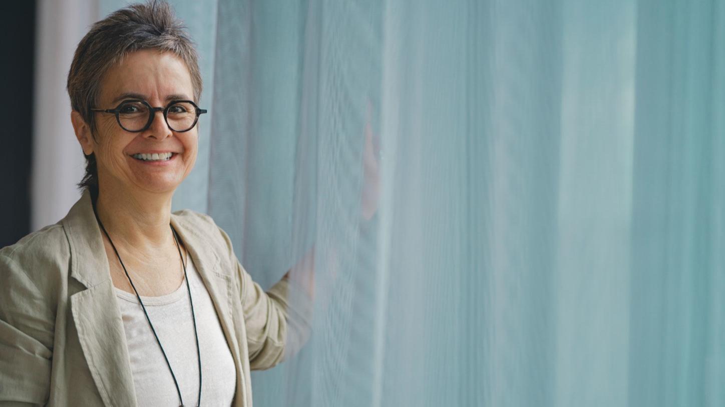 Portrait photo of Stefanie Vonhoff. She is in her mid-50s, wears short gray hair, a beige blazer, a striking necklace and black glasses and can be seen up to her upper body. She stands in front of a window with a white curtain and smiles at the camera.